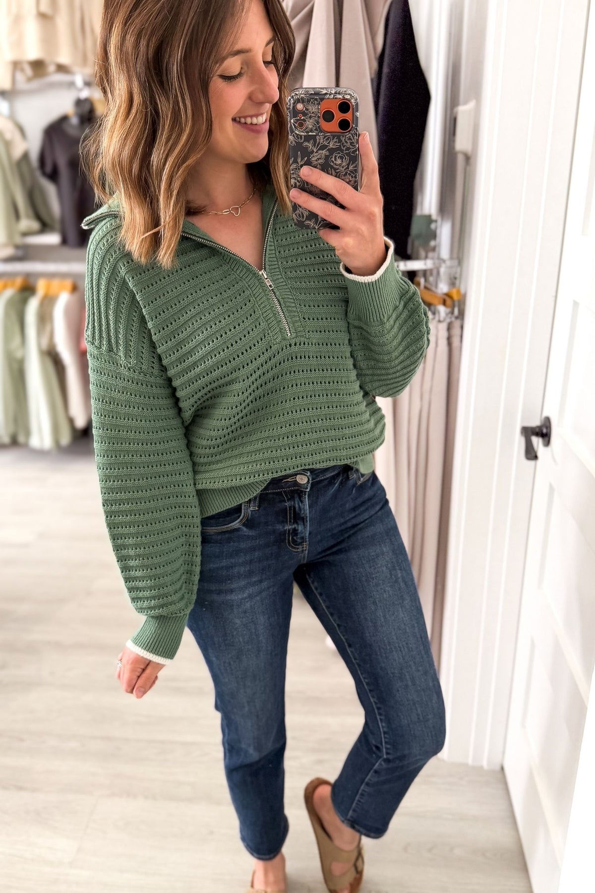 A woman takes a mirror selfie in a boutique while wearing a textured sage green half-zip pullover sweater, dark wash blue jeans, and tan flat shoes.