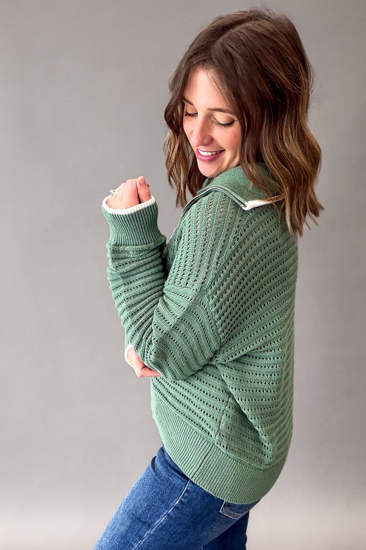 A woman wearing a sage green textured knit zip-up hoodie sweater and blue jeans, posing against a neutral gray studio background.
