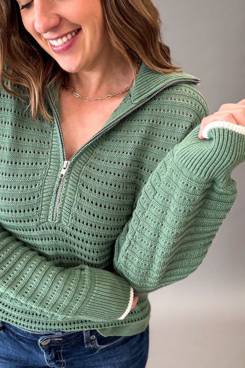 A woman wearing a sage green knit half-zip sweater with textured openwork patterns and ribbed cuffs, paired with blue jeans against a neutral gray background.