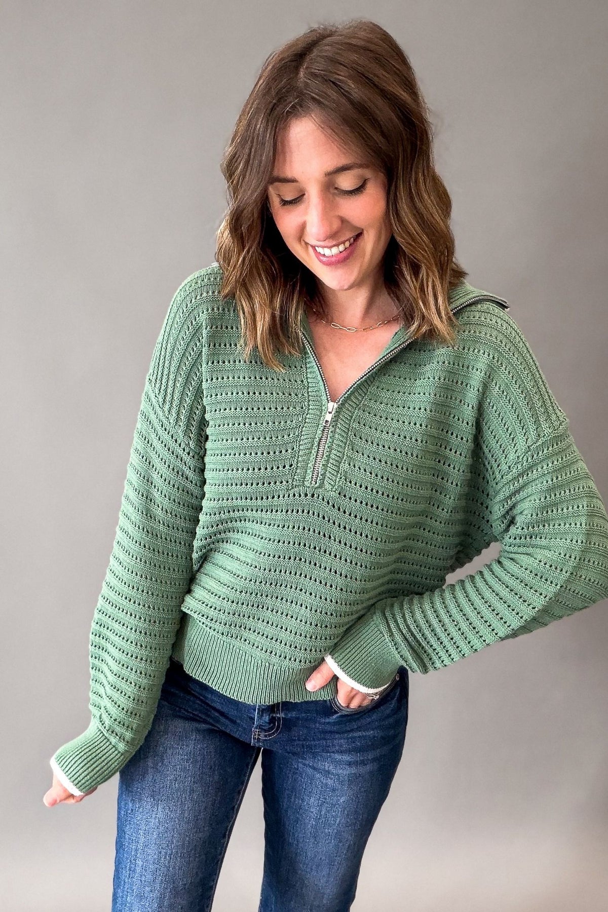 A woman wearing a sage green textured knit half-zip pullover sweater and blue jeans, standing against a neutral gray studio background.