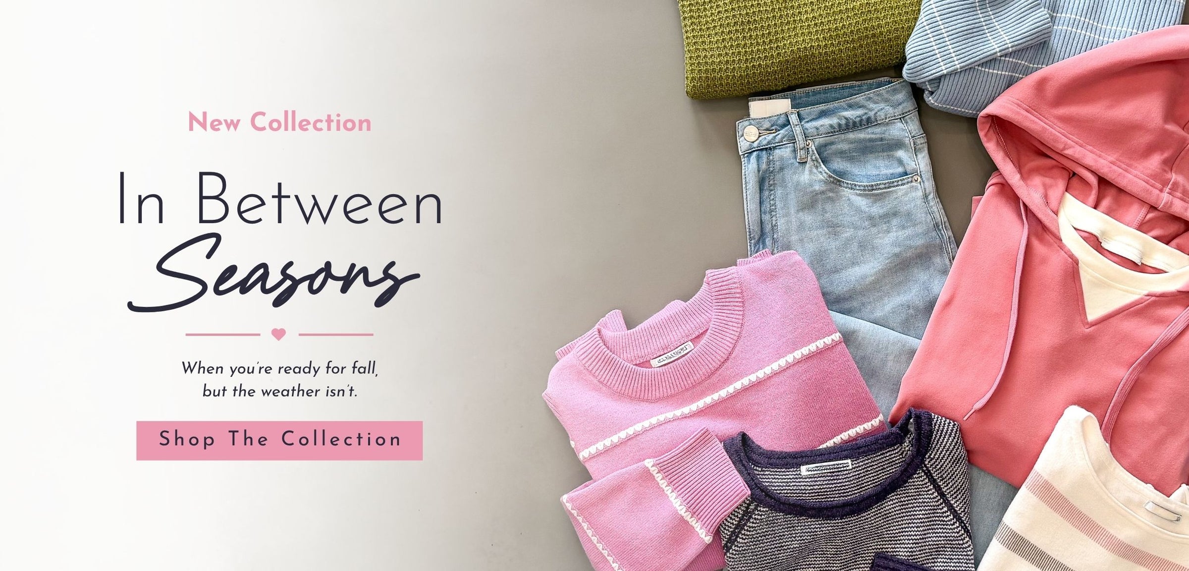A promotional flat lay showing a folded collection of women's clothing on a light neutral background, with text reading 'New Collection In Between Seasons' and 'Shop The Collection.' Visible items include pink and coral hoodies, light blue jeans, a pink knit top, a gray top, and a striped cream garment, plus a small olive-green folded item at the top.