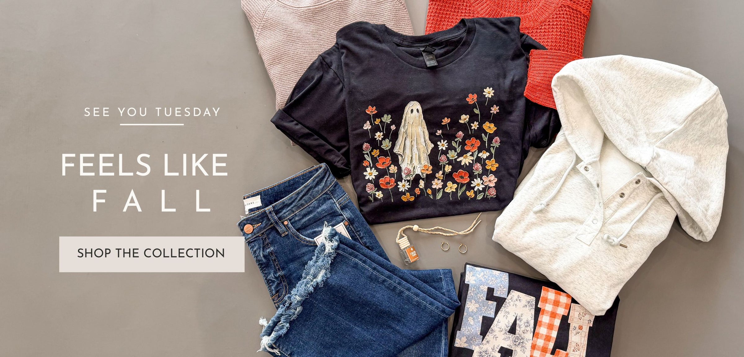 A styled flat-lay of fall clothing on a taupe background, featuring a black graphic T-shirt with a ghost and floral print, blue jeans, a cream button-up shirt, and other coordinating apparel. Text on the left reads 'SEE YOU TUESDAY', 'FEELS LIKE FALL', and 'SHOP THE COLLECTION'.