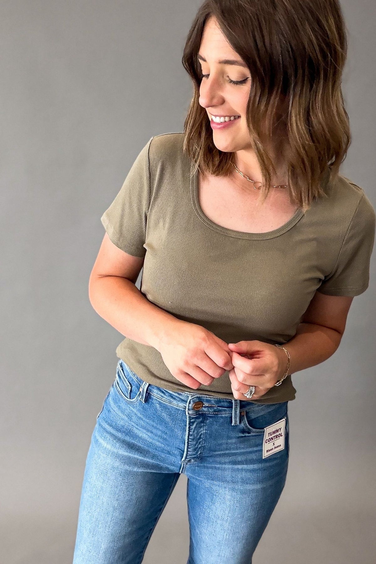A woman wearing a short-sleeve scoop-neck t-shirt in olive green and blue jeans, standing against a plain gray studio background.