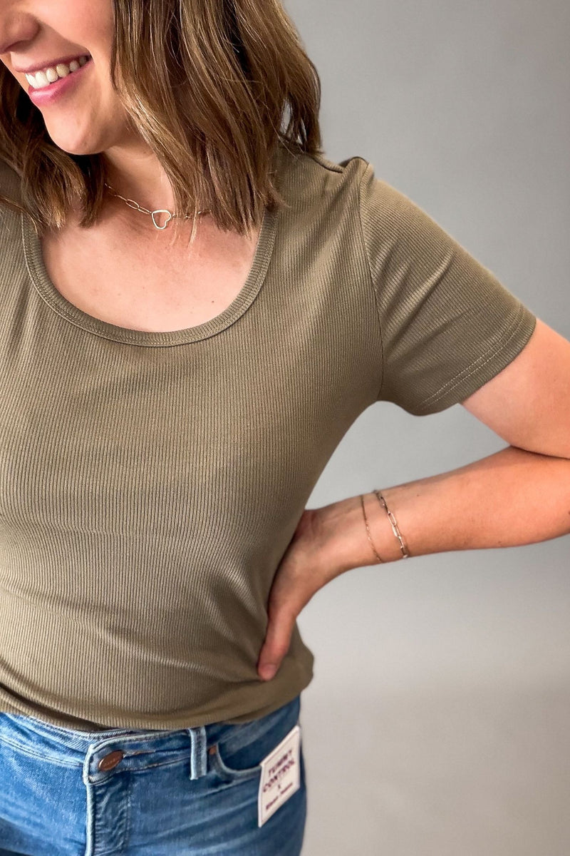 A woman wearing a short-sleeve ribbed scoop-neck t-shirt in olive green, paired with blue jeans, shown from the side against a plain gray background.