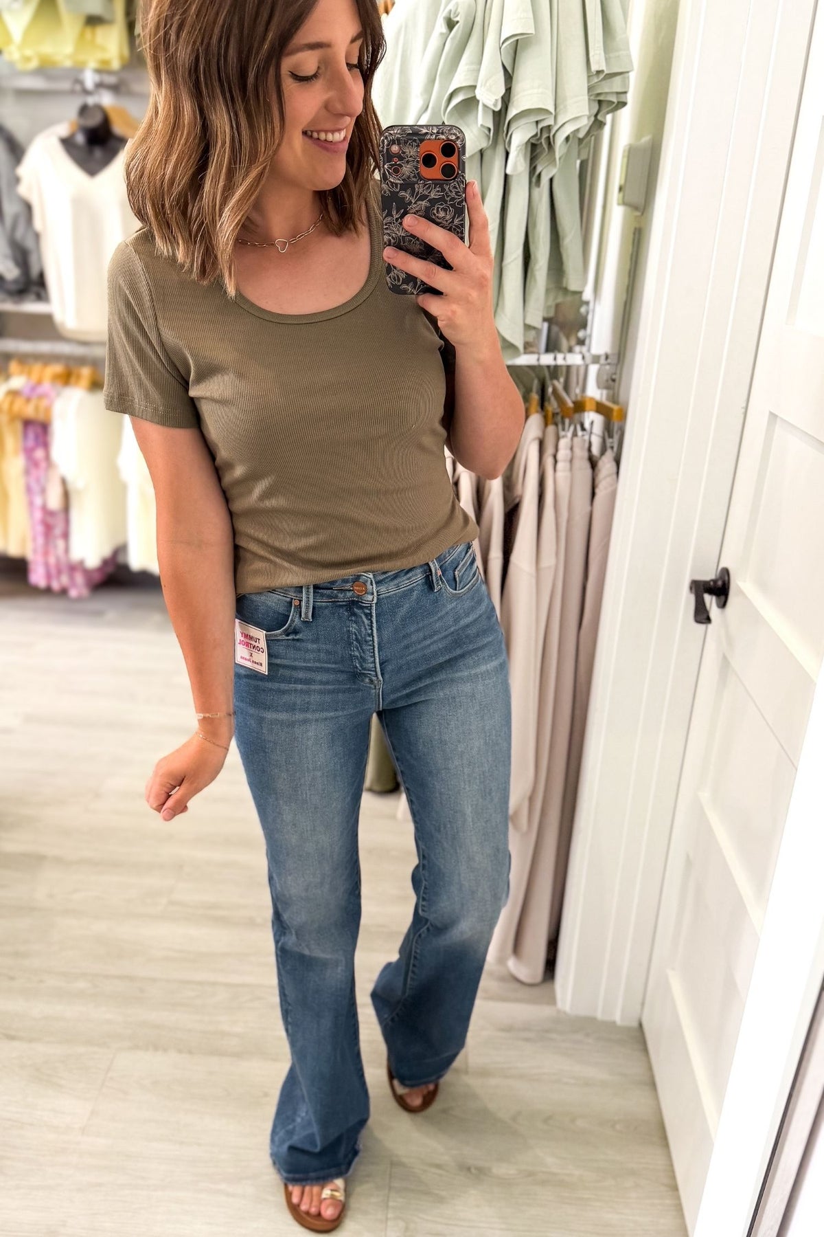 A woman takes a mirror selfie in a clothing store, wearing a short-sleeve olive green ribbed t-shirt and blue flare jeans.