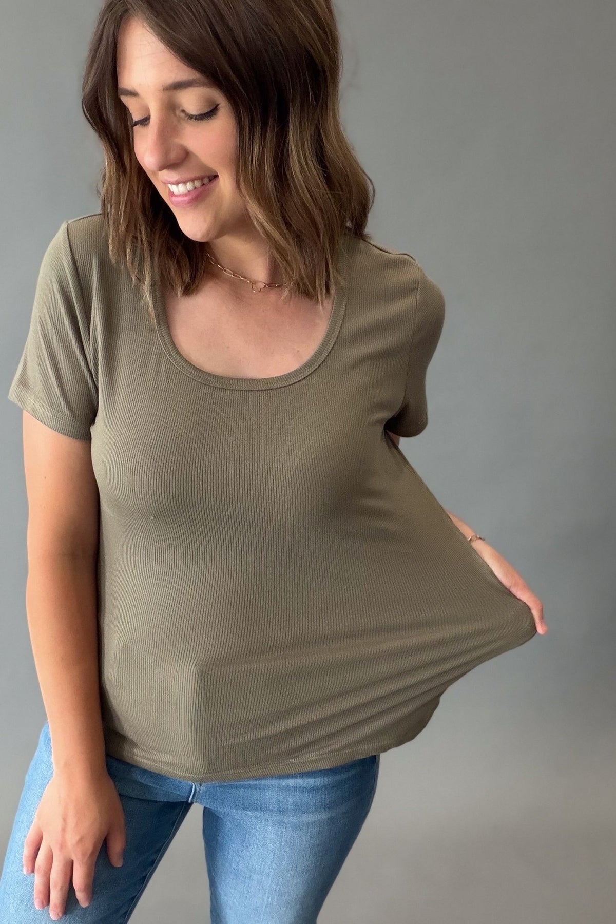 A woman wearing a short-sleeve scoop-neck ribbed t-shirt in olive green, holding out her left side to demonstrate the fabric's stretch, paired with blue jeans against a solid gray background.
