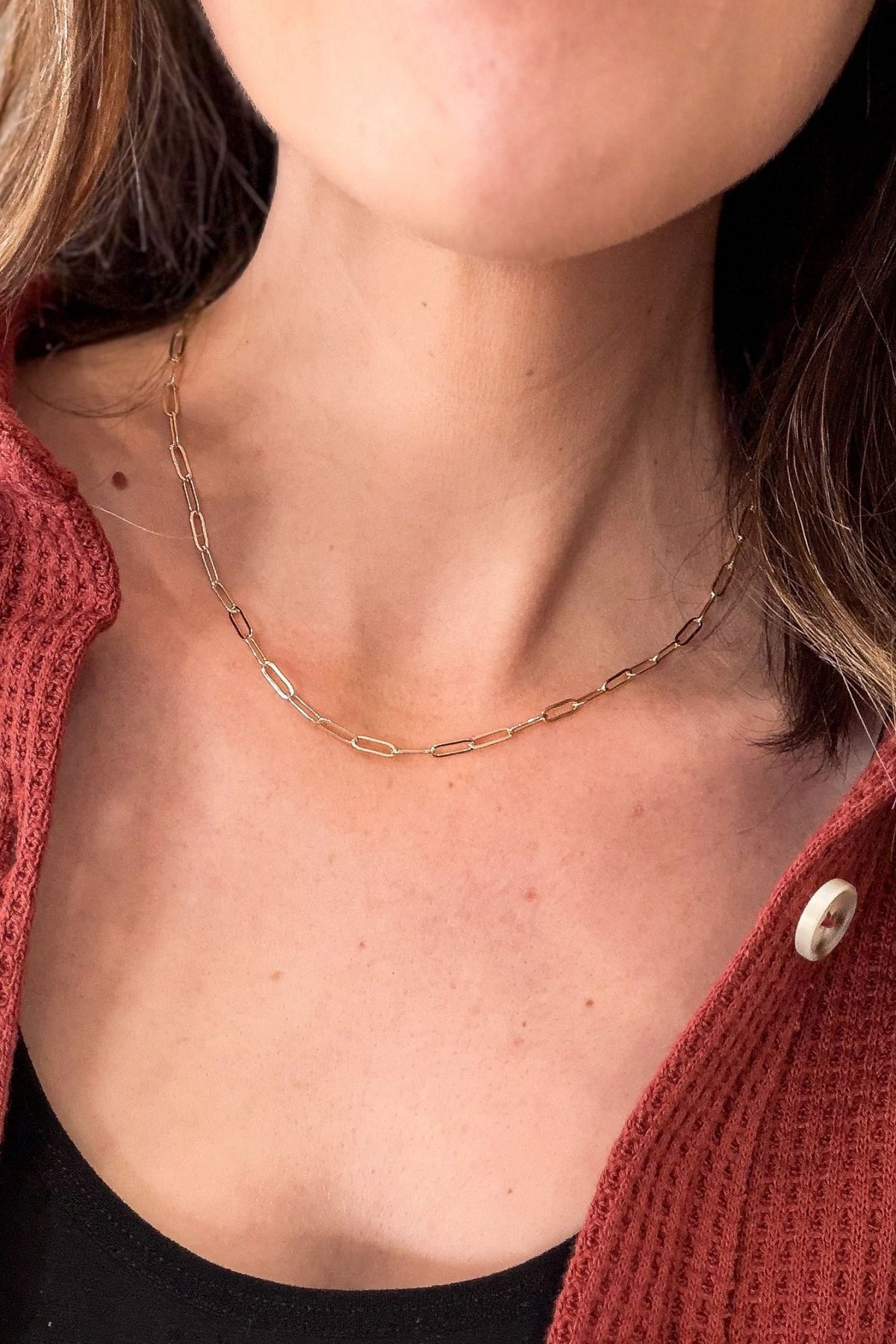 A close-up of a model wearing a delicate gold paperclip chain necklace, styled with a rust-colored knit cardigan and a black top.