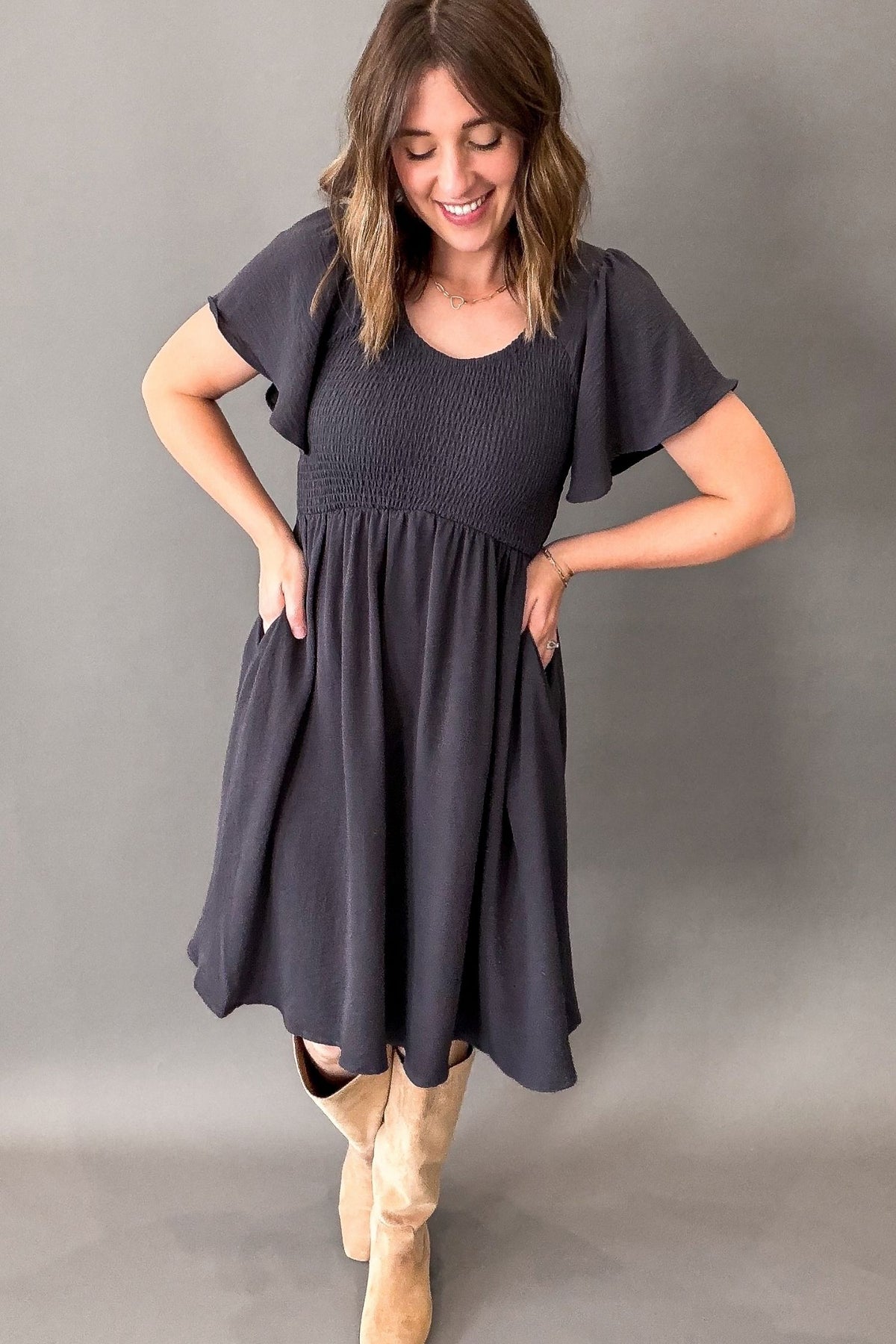 woman wearing a smoky blue smocked bust dress with flutter sleeves, flowy skirt and tall neutral colored boots against a grey background.