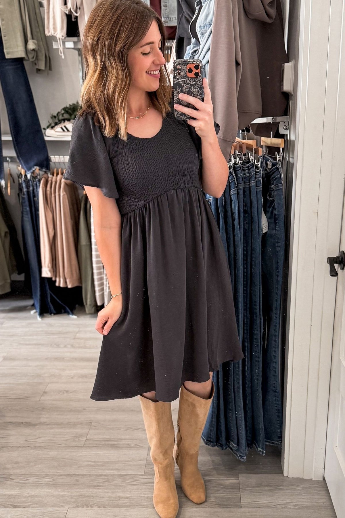 A woman takes a mirror selfie in a clothing boutique wearing a dark charcoal blue/gray short-sleeve midi dress and tan knee-high suede boots. Clothing racks and jeans are visible in the background.