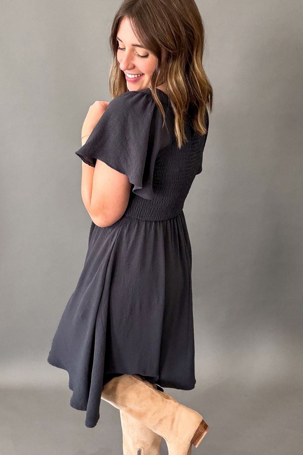 A woman wearing a dark blue/charcoal gray dress with flutter sleeves and a smocked bodice, paired with beige boots, photographed against a neutral gray studio background.