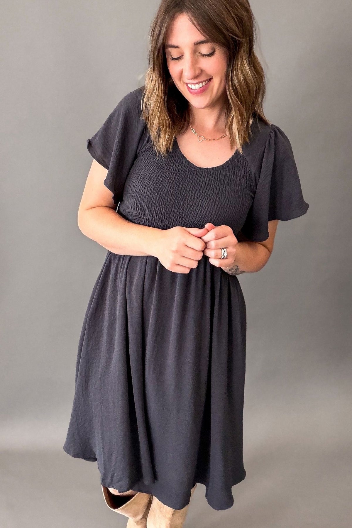 A woman wearing a dark blue/charcoal gray smocked dress with flutter sleeves, standing against a neutral gray studio background.