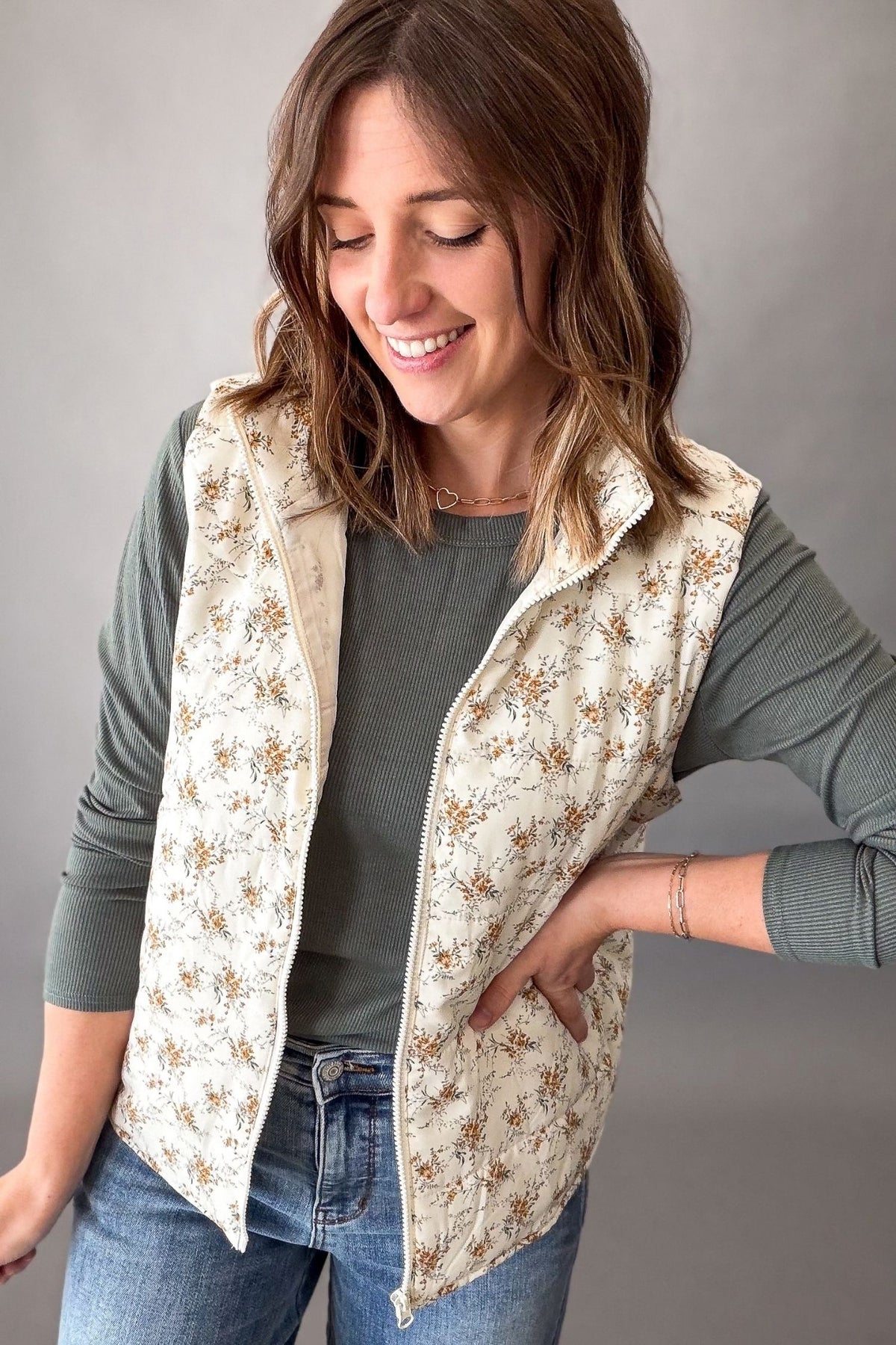 A woman wearing a cream-colored quilted vest with a brown floral pattern over a dark green long-sleeve shirt and blue jeans, posing against a neutral gray studio background.