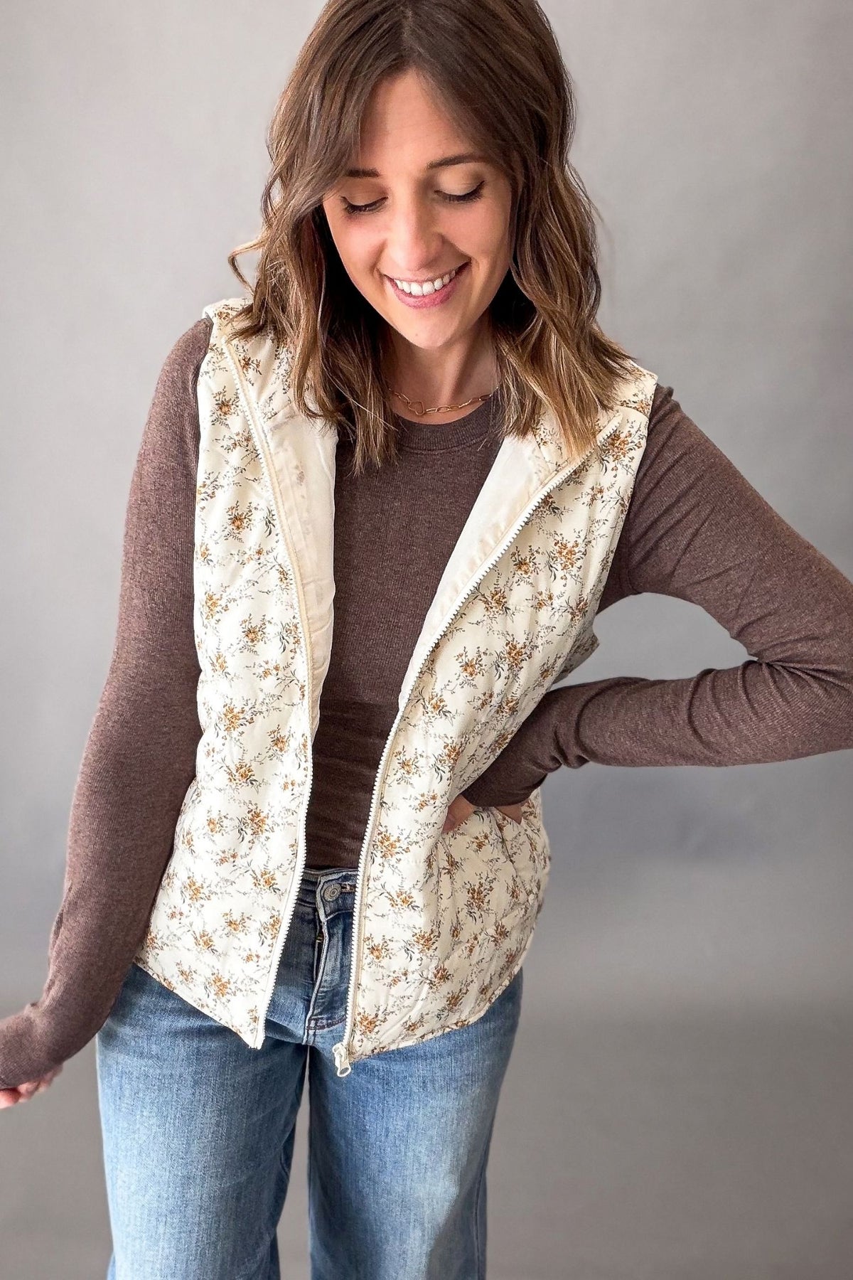 A woman wearing a sleeveless quilted vest with a beige and brown floral pattern over a brown long-sleeve shirt and blue jeans, standing against a plain gray studio background.