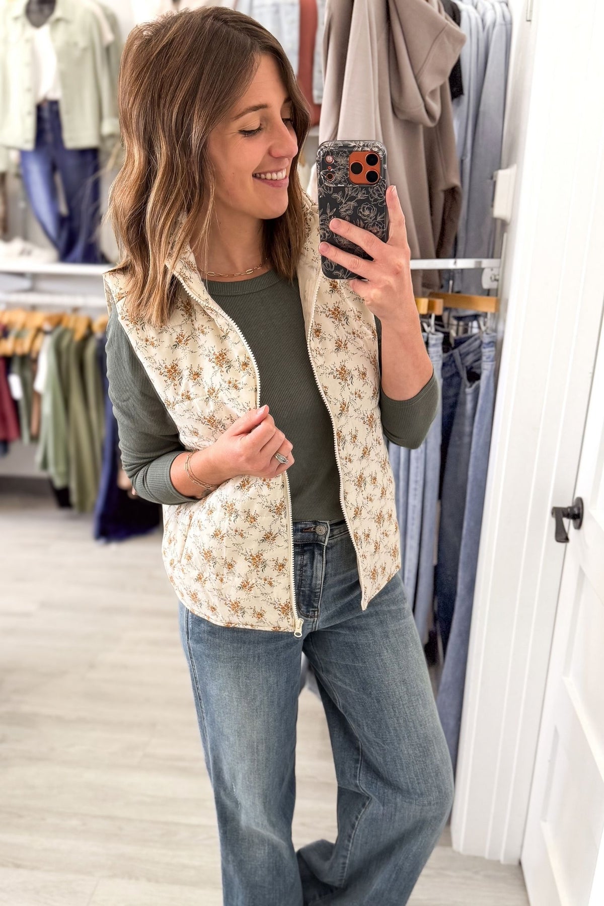 A woman takes a mirror selfie in a clothing store while wearing a cream and brown floral print quilted vest over an olive green long-sleeve shirt and blue jeans.