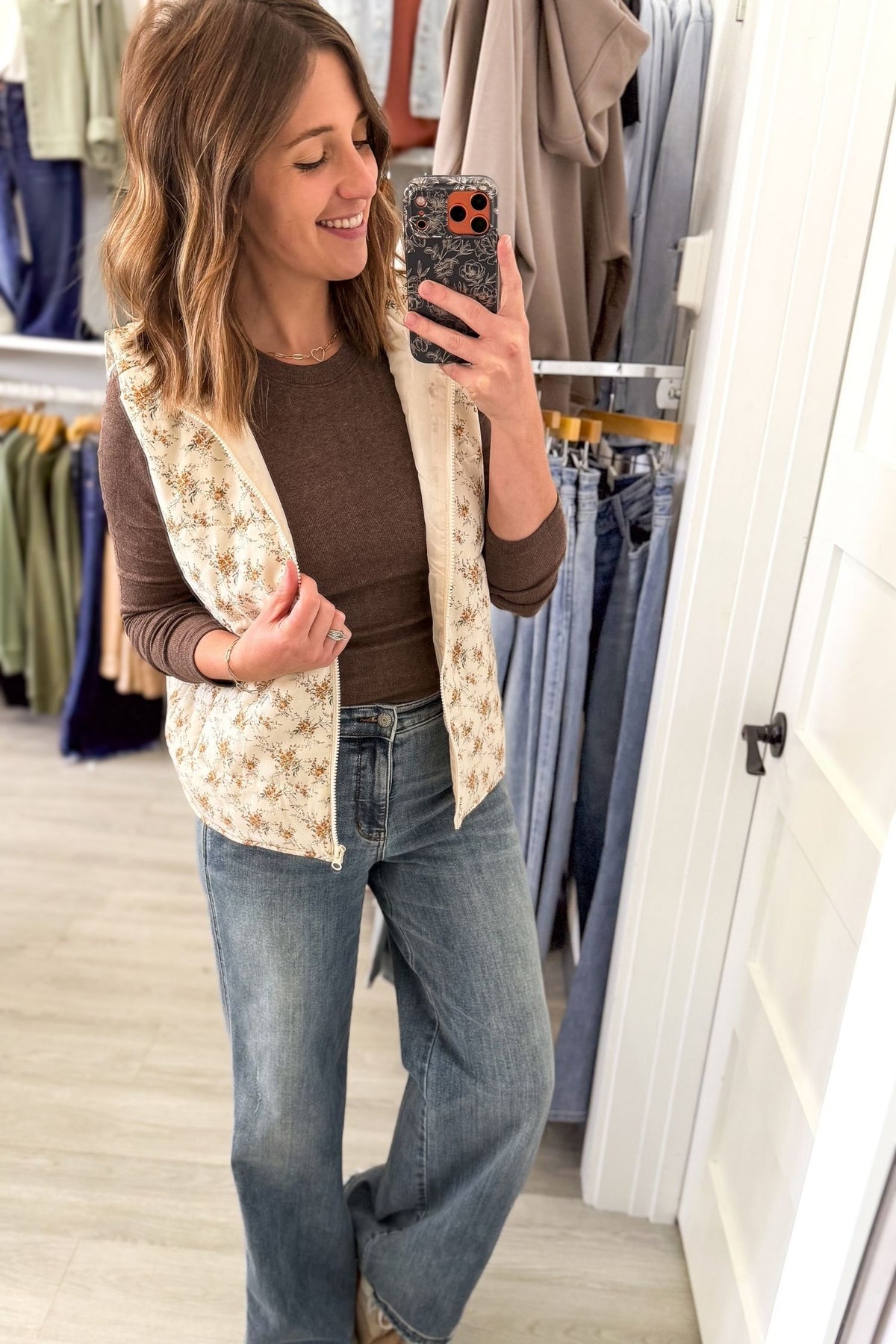 A woman takes a mirror selfie in a boutique while wearing a cream floral quilted vest over a brown long-sleeve top and blue jeans.