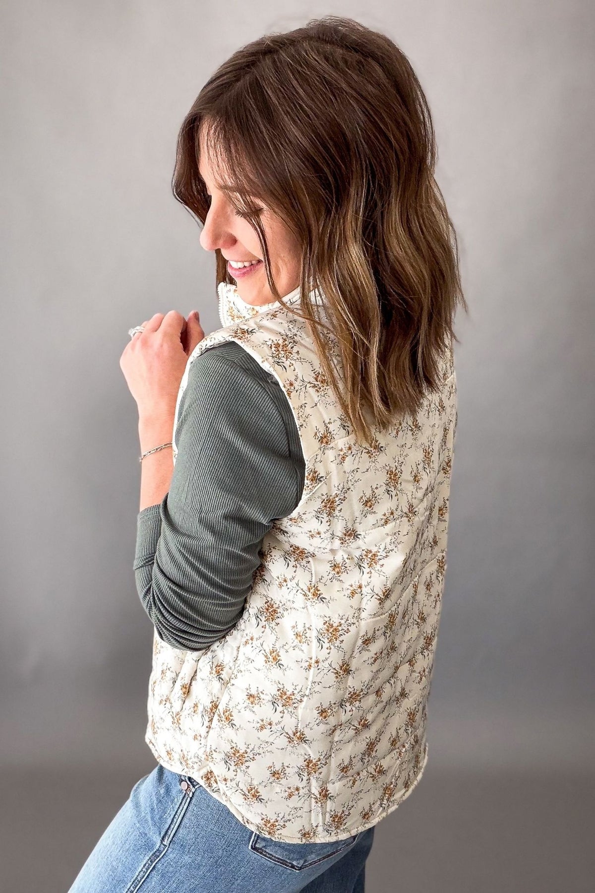 A woman wearing a sleeveless vest with a beige and brown floral pattern over a dark green long-sleeve shirt and blue jeans, photographed against a neutral gray studio background.