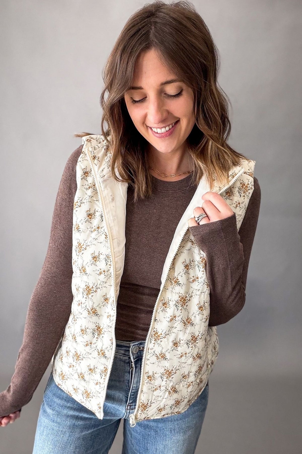 A woman wearing a cream-colored vest with a brown floral pattern over a brown long-sleeve shirt and blue jeans, standing against a neutral gray background.
