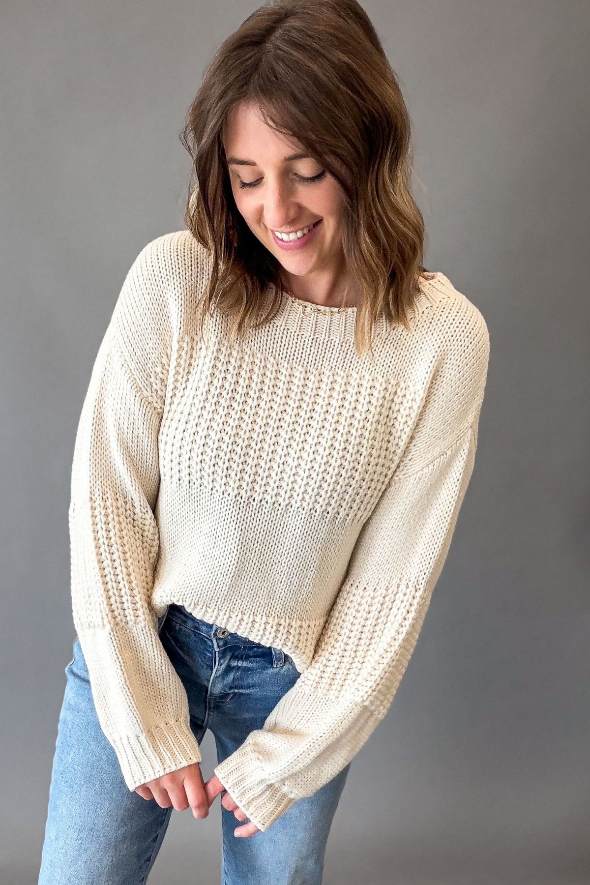 A woman wearing a ivory-colored, textured knit sweater and blue jeans, posing against a solid gray studio background.