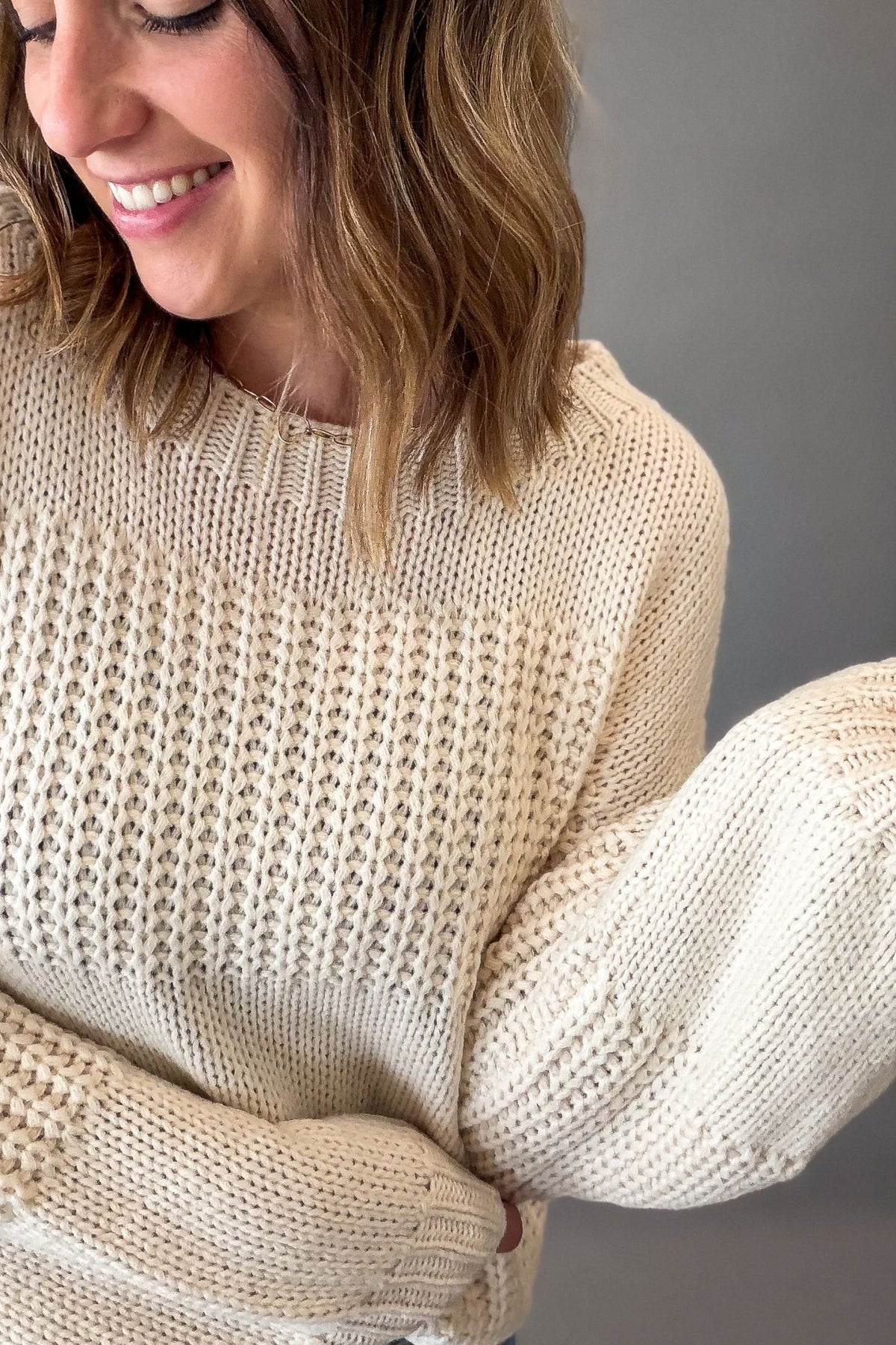 A woman wearing a chunky knit ivory-colored sweater with textured vertical panels and balloon sleeves, shown in a close-up shot against a neutral gray background.