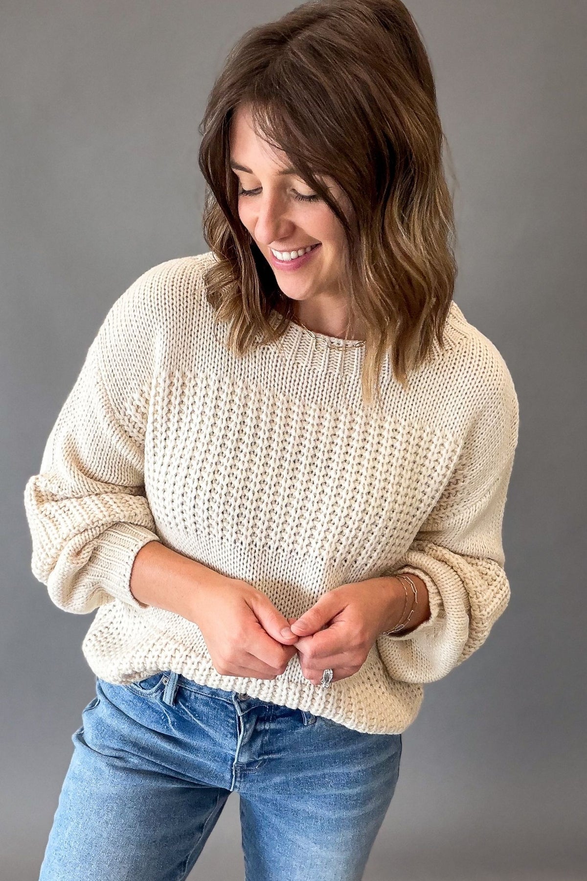 A woman wearing a ivory-colored chunky knit sweater and blue jeans, looking down and smiling against a solid gray studio background.