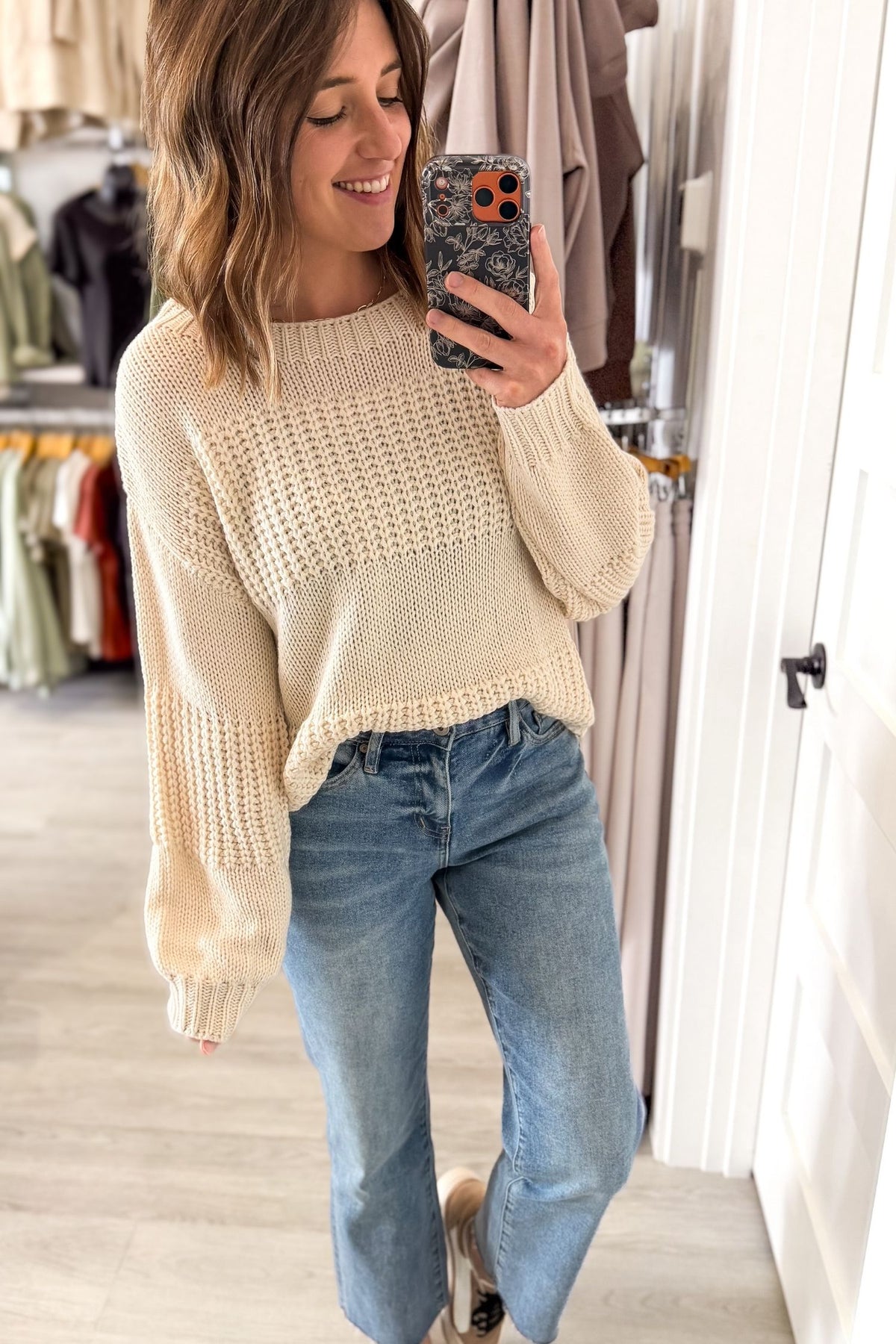 A woman takes a mirror selfie in a boutique while wearing a chunky knit ivory sweater and blue jeans.