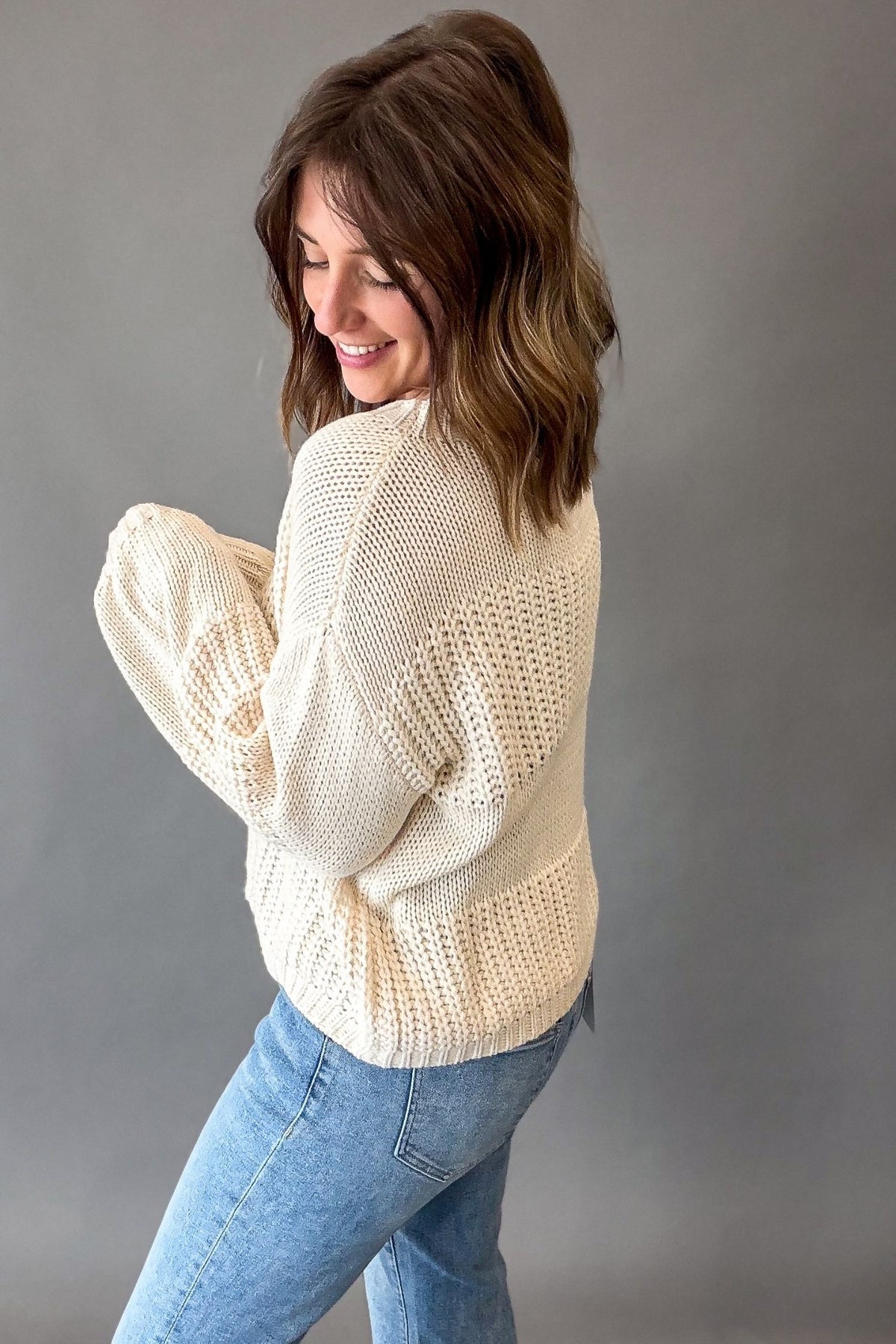 A woman wearing a ivory-colored, open-knit sweater and light blue jeans, photographed from a side-back angle against a solid gray studio background.