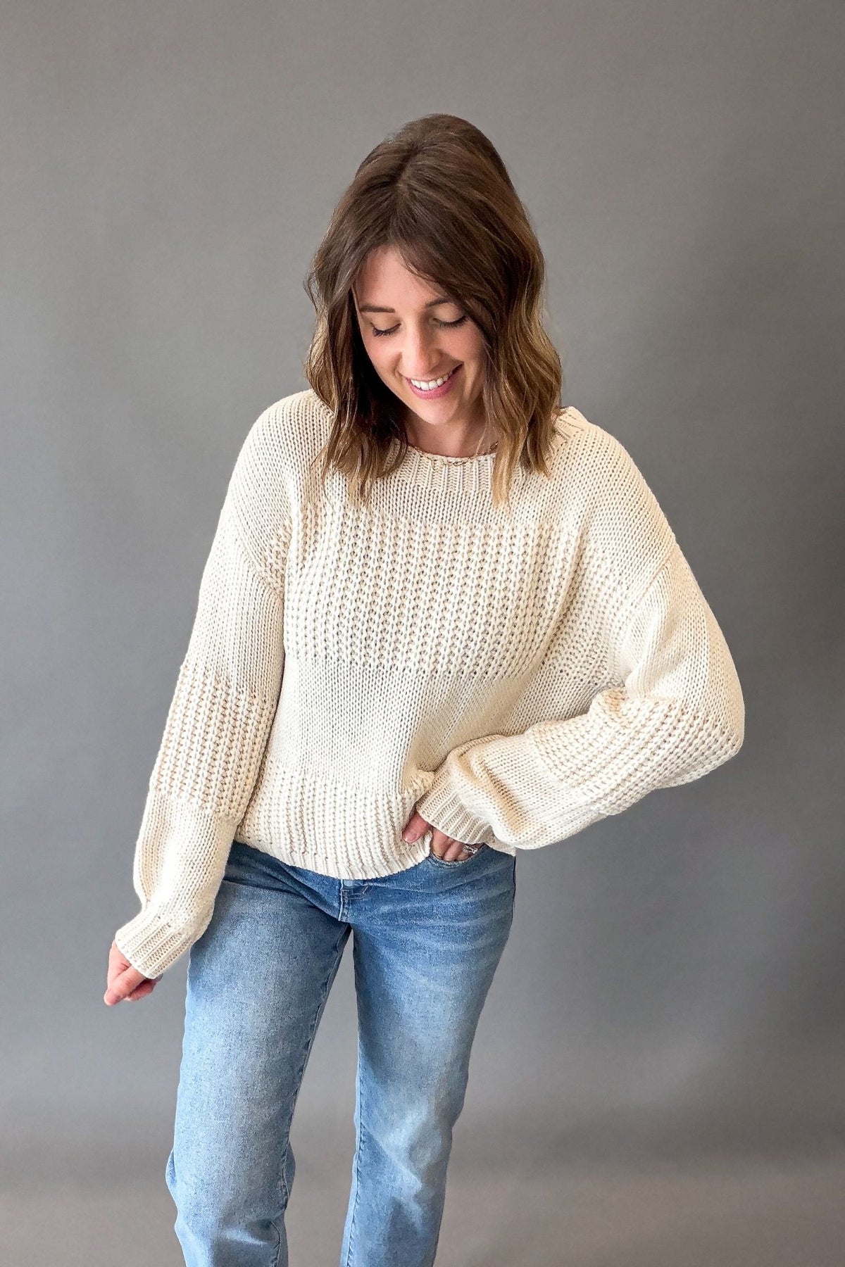A woman wearing a ivory-colored textured knit sweater and blue jeans, standing against a solid gray studio background.