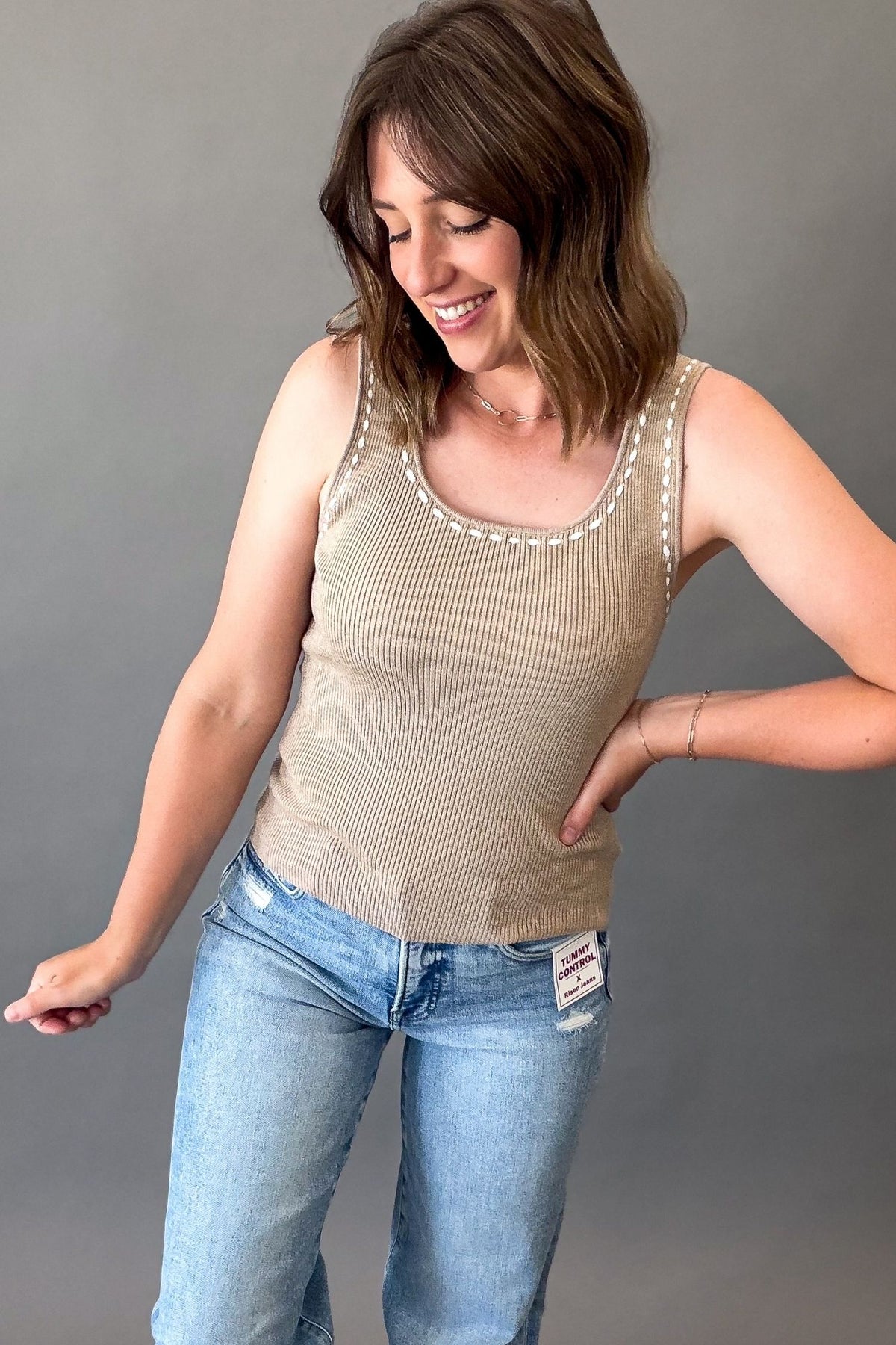 A woman wearing a beige ribbed knit tank top with off white trim along the neckline, paired with light blue jeans, posing against a solid gray background.