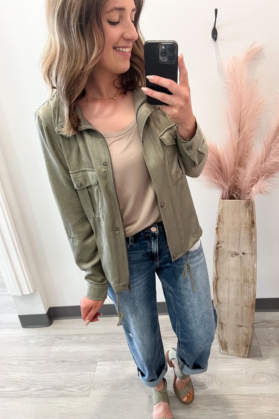 Woman taking a mirror selfie wearing an olive green jacket, beige top, and blue jeans.