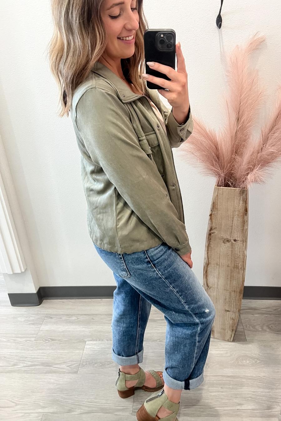 Woman taking a selfie wearing an olive green jacket and blue jeans, standing next to a decorative plant.