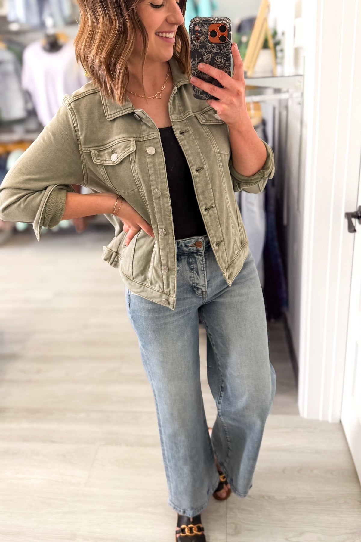 Woman taking a mirror selfie wearing an olive green jacket, black top, and light blue jeans.