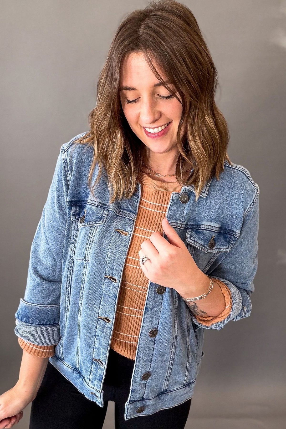 A woman wearing a light-wash blue denim jacket over a rust-orange and white striped knit top, paired with black pants, photographed against a neutral gray studio background.