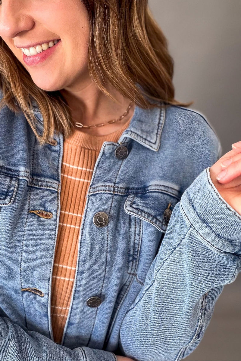 A close-up of a woman wearing a light blue denim jacket over a rust-orange ribbed knit top, with a neutral gray background.