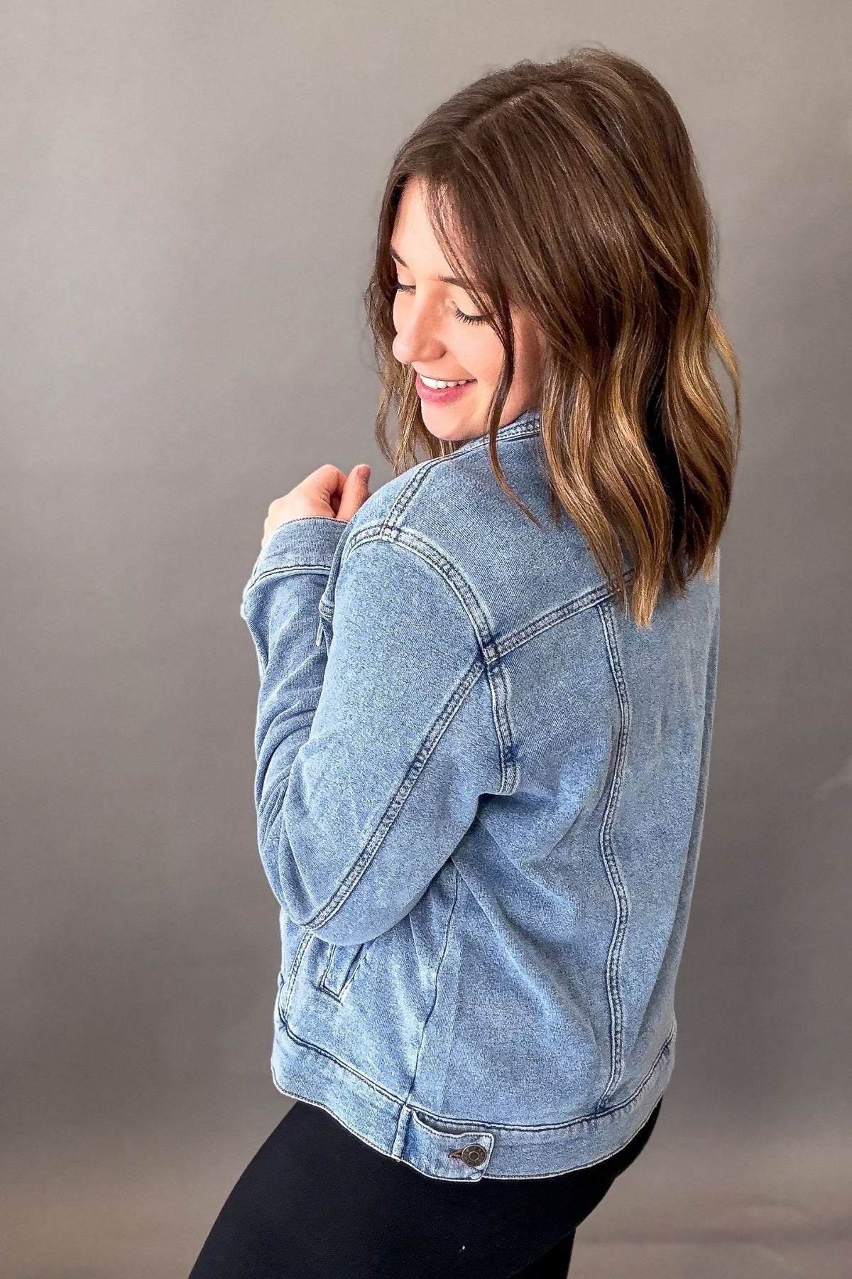 A woman with shoulder-length brown hair is shown from a side-back angle wearing a light-wash blue denim jacket and black pants against a plain gray studio background.