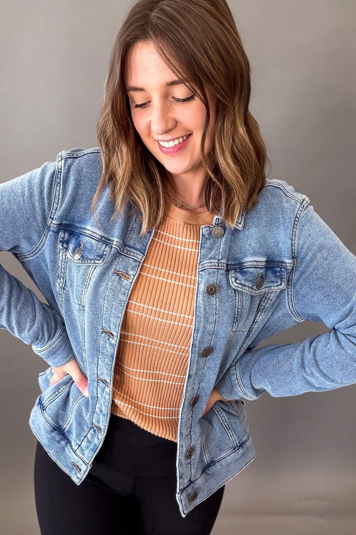 A woman wearing a light blue denim jacket over a tan and white striped knit top, paired with black pants, posing against a neutral gray studio background.