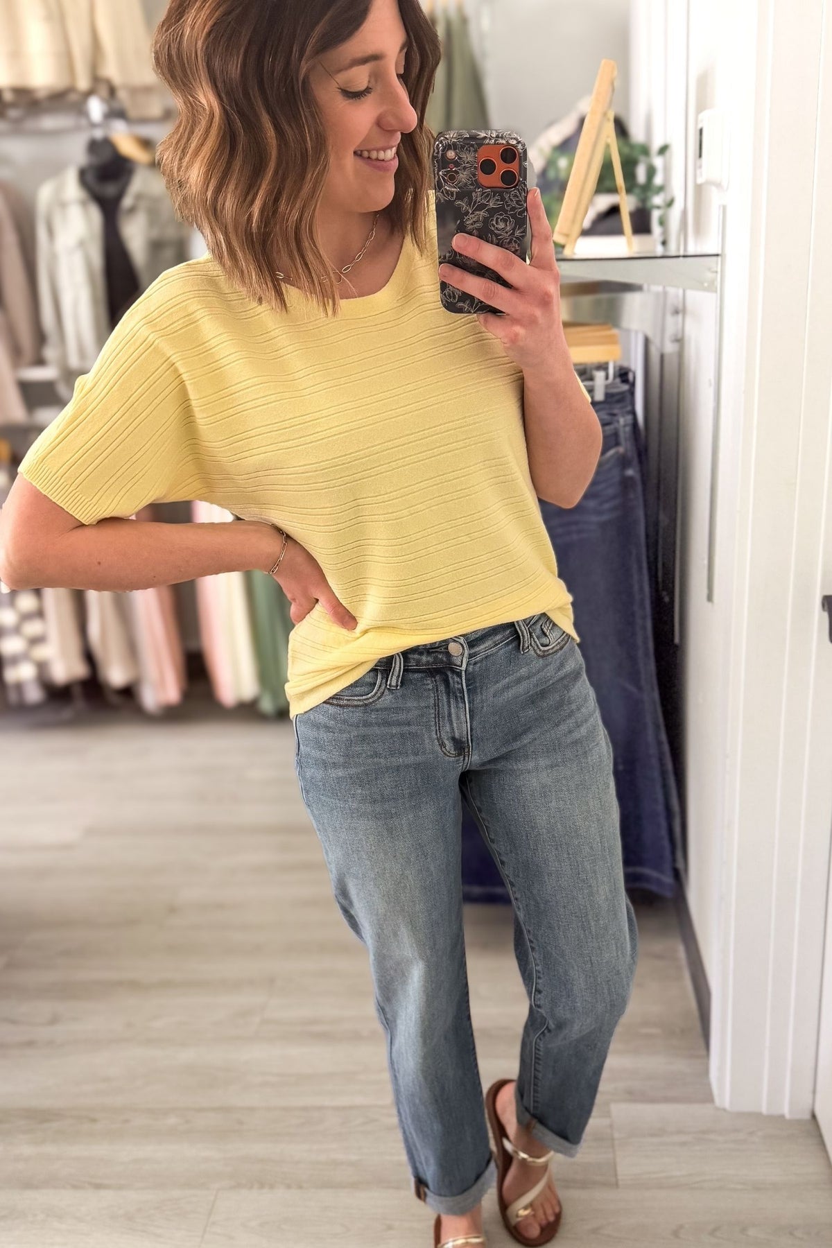 Woman taking a mirror selfie wearing a yellow top and blue jeans in a clothing store.