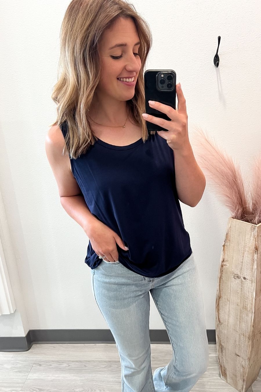 Woman taking a mirror selfie wearing a navy top and light blue jeans, standing against a white wall with decorative elements.