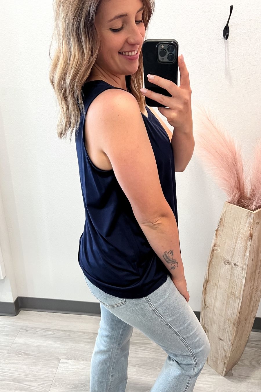 Woman taking a mirror selfie wearing a navy sleeveless top and light blue jeans.