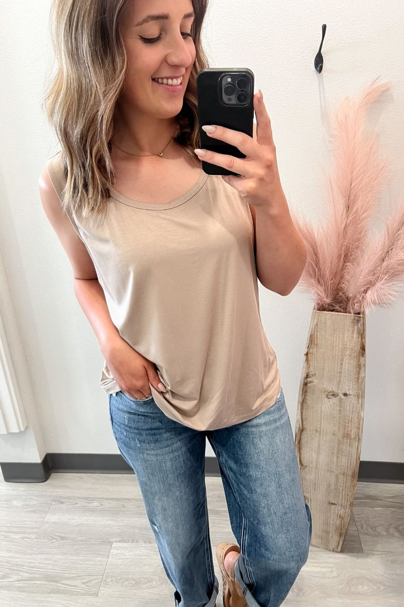 Woman taking a mirror selfie wearing a beige tank top and blue jeans, holding a phone.