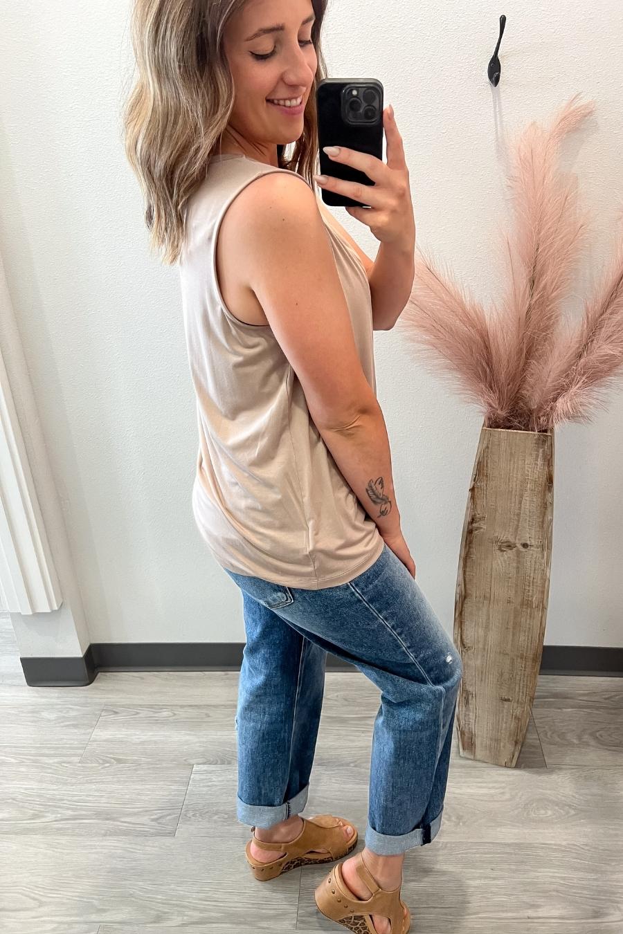 Woman taking a mirror selfie wearing a beige tank top and blue jeans, standing next to a vase with pink decorative grasses.