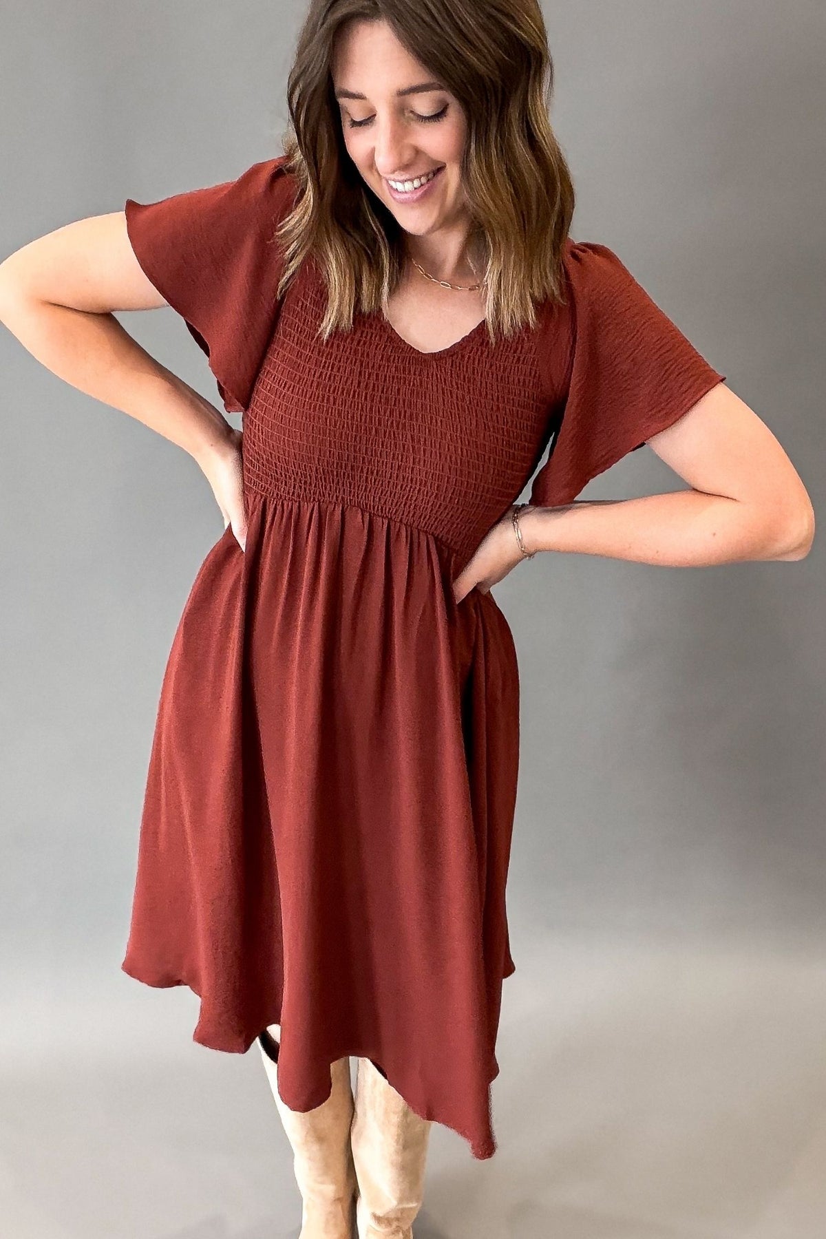 A woman modeling a rust-colored short-sleeve dress with a smocked bodice and flared skirt, standing against a plain gray studio background.