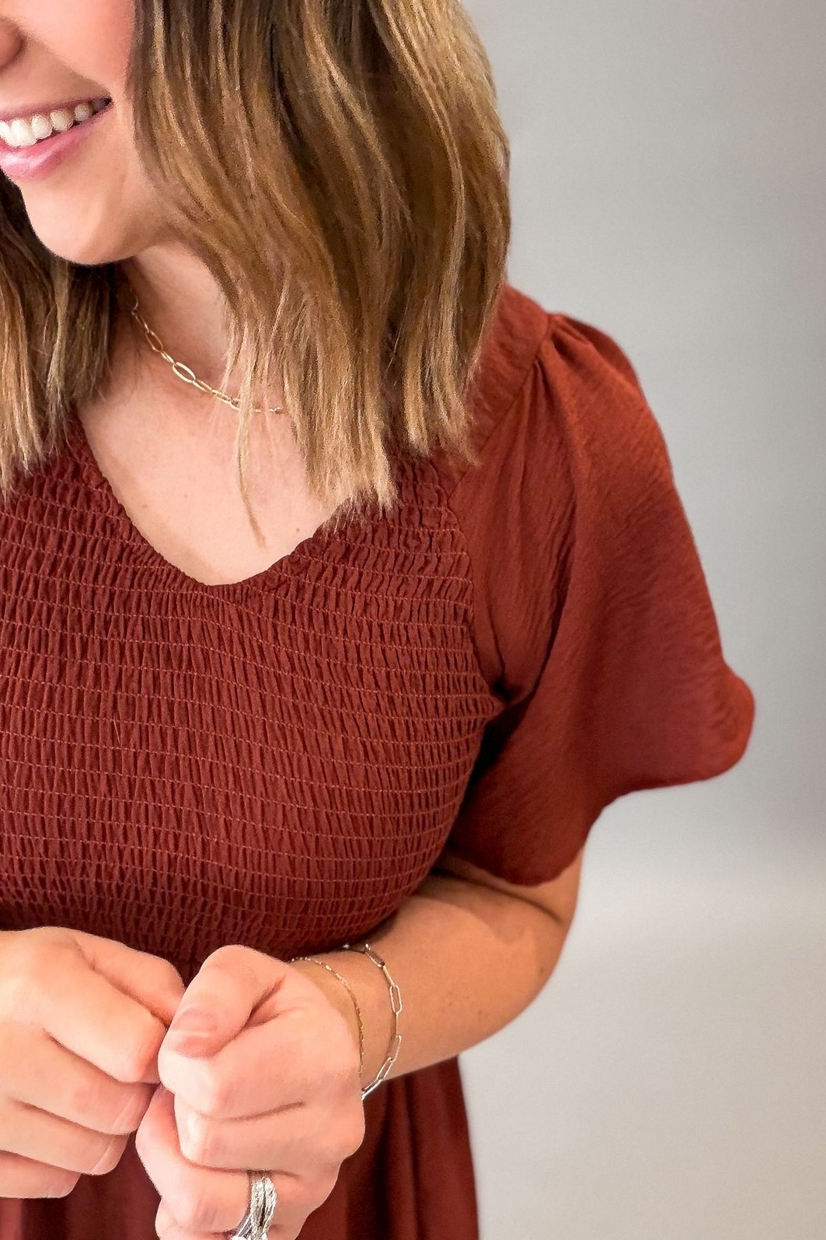 A close-up of a woman wearing a rust-brown smocked short-sleeve top, showing her hands, jewelry, and hair against a neutral gray background.