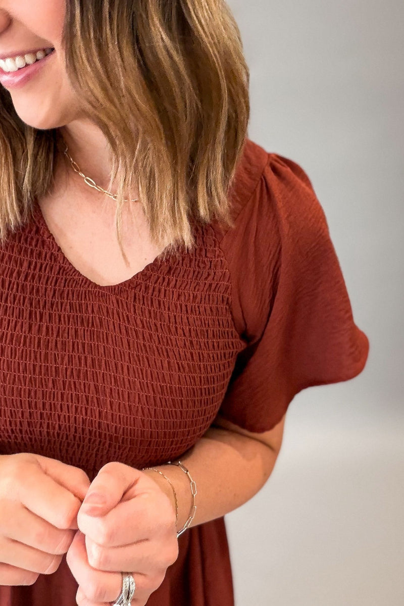 A close-up of a woman wearing a rust-brown smocked short-sleeve top, showing her hands, jewelry, and hair against a neutral gray background.