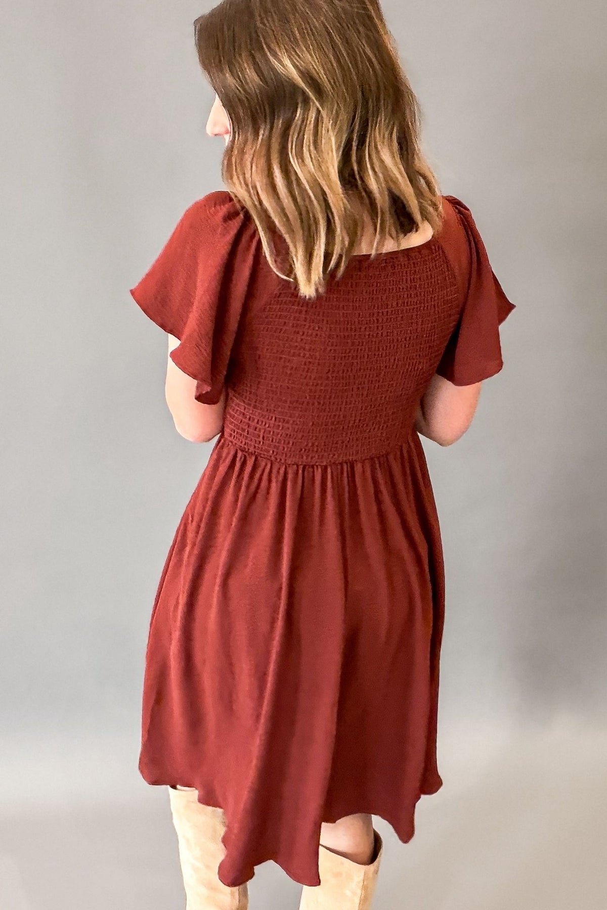 A woman shown from the back wearing a rust-colored smocked dress with flutter sleeves and tan boots, standing against a neutral gray studio background.