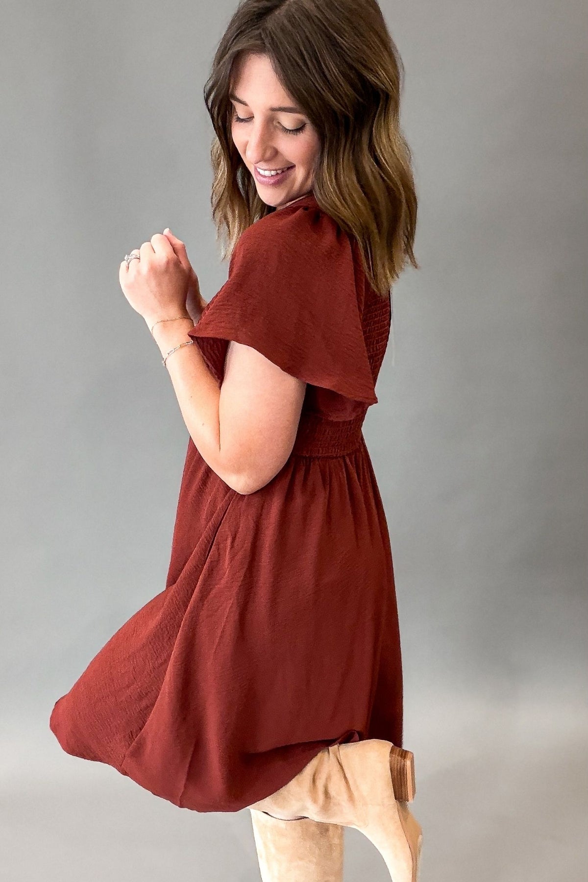 A woman wearing a rust-colored short-sleeve dress with flutter sleeves and a gathered waist, paired with beige boots, posing against a neutral gray studio background.
