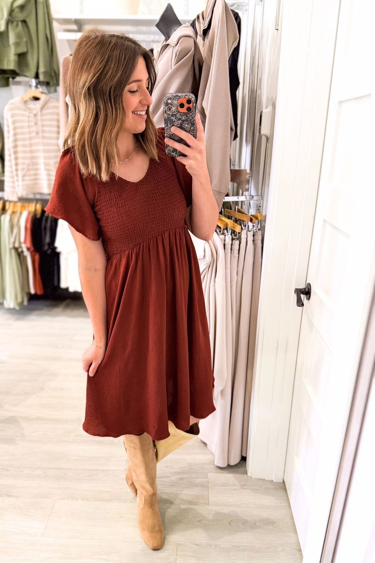 A woman takes a mirror selfie in a clothing boutique wearing a rust-brown smocked short-sleeve dress and tan ankle boots. Clothing racks and a white door are visible in the background.