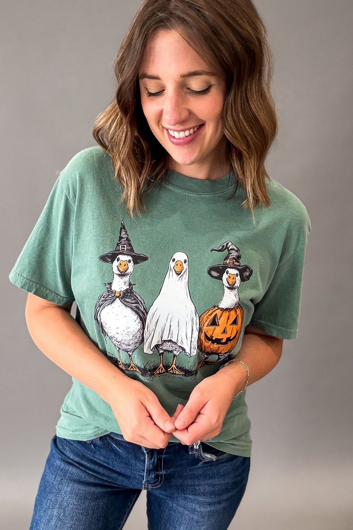 A woman wearing a sage green graphic t-shirt with a cartoon illustration of three geese dressed in Halloween costumes (witch hat, ghost, pumpkin), paired with blue jeans, against a plain gray studio background.