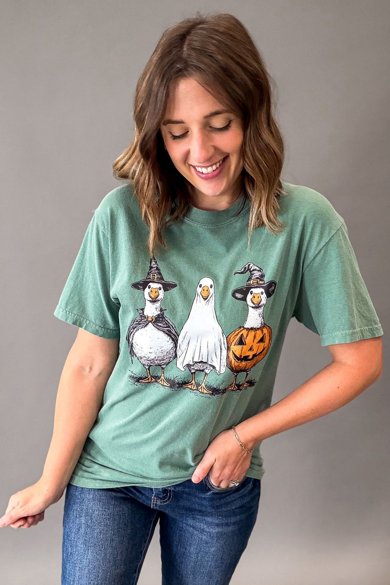 A woman wearing a sage green graphic t-shirt with a cartoon illustration of three geese dressed in Halloween costumes (witch hats, ghost, and jack-o'-lantern), paired with blue jeans, against a solid gray studio background.