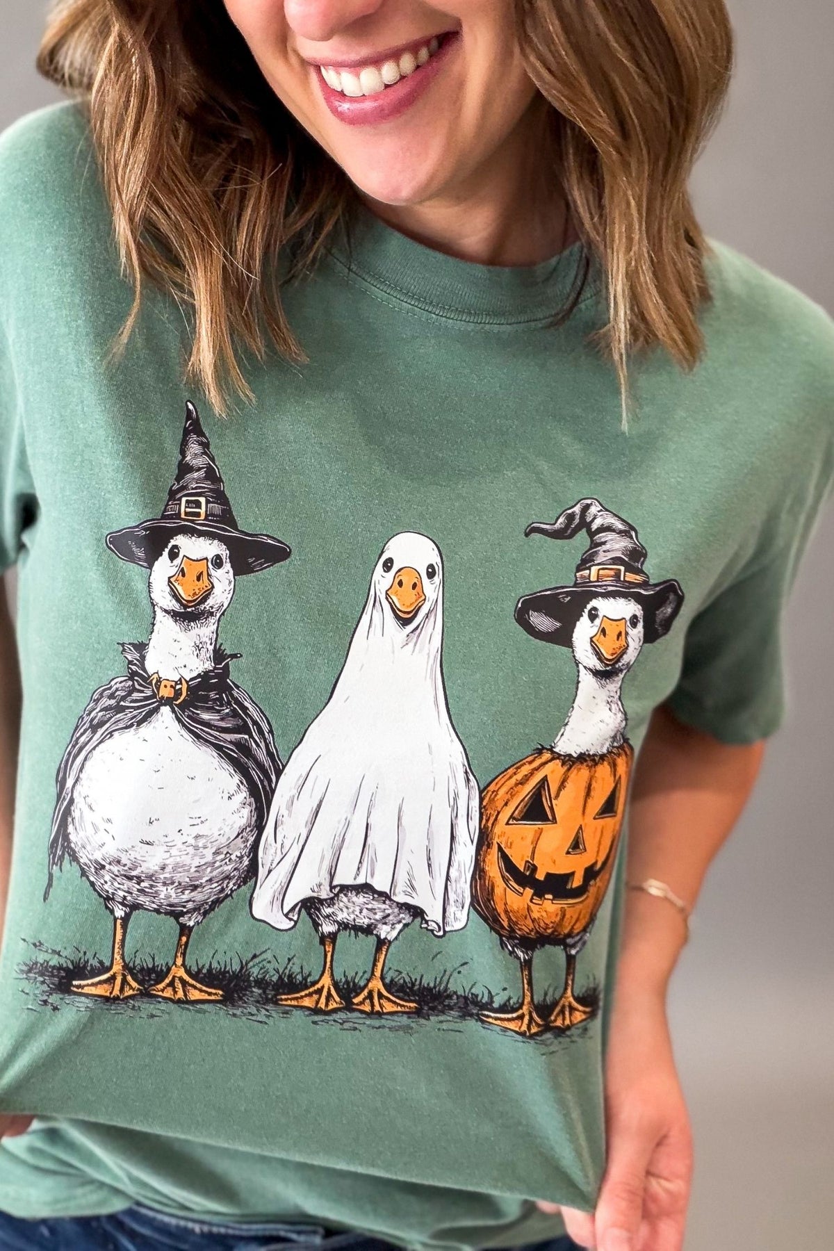A woman wearing a sage green graphic t-shirt featuring three geese dressed as Halloween: a witch, a ghost, and a jack-o'-lantern.