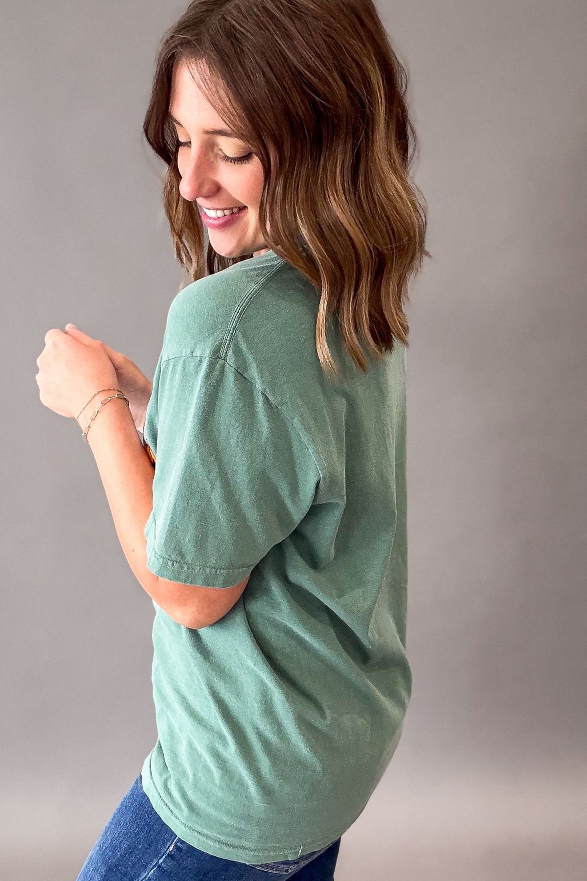 A woman with shoulder-length brown hair is shown from the back wearing an oversized sage green t-shirt and blue jeans, posing against a neutral gray studio background.